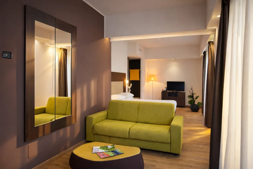 Nell'immagine, l'Executive Suite dell'Hotel Palace Lucera: si vede in primo piano la zona giorno, con un tavolino basso e un divano di colore verde. Sullo sfondo, alle spalle del divano, si vede il letto matrimoniale.