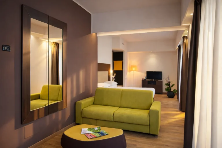 Nell'immagine, l'Executive Suite dell'Hotel Palace Lucera: si vede in primo piano la zona giorno, con un tavolino basso e un divano di colore verde. Sullo sfondo, alle spalle del divano, si vede il letto matrimoniale.