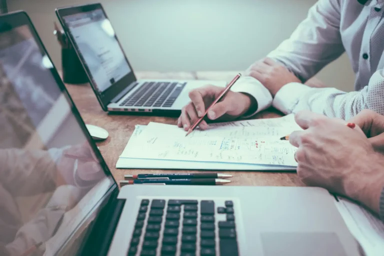 Nell'immagine, si vedono le mani di due persone impegnate a lavorare ai loro laptop. Sulla scrivania ci sono anche dei fogli con degli appunti.