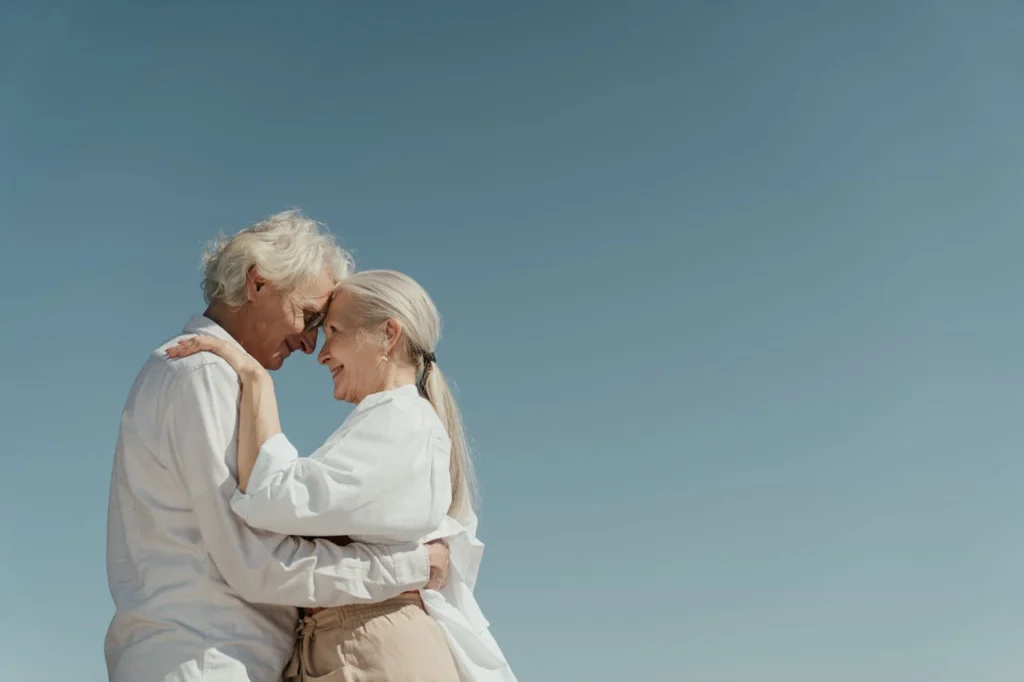 Nell'immagine, una coppia di anziani abbracciati che sorridono guardandosi negli occhi.